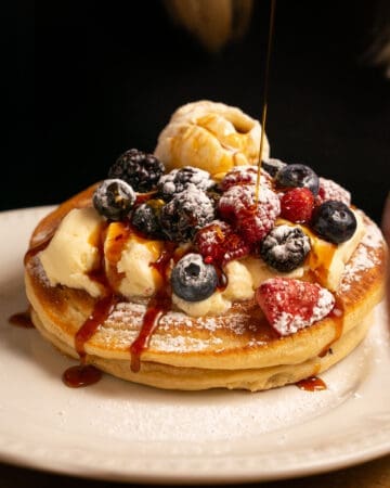 syrup being poured over stack of pancakes with strawberries blueberries and ice cream